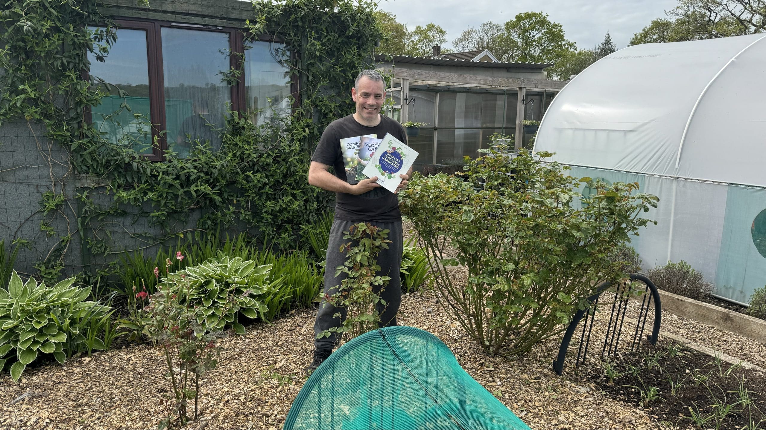 Tony O'Neill standing in his productive South Wales garden holding all three of his vegetable growing books — Composting Masterclass, Your First Vegetable Garden, and Simplify Vegetable Gardening — with the polytunnel and lush ornamental planting visible behind him