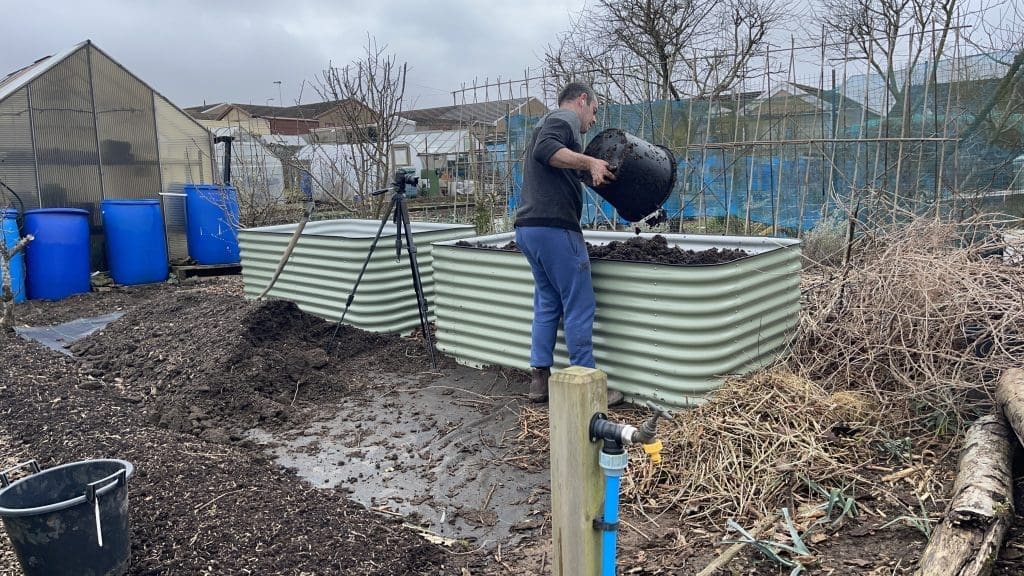 Tony O'Neill tipping a large bag of hugelkulture ingredients into one of two tall Birdies raised garden beds at his allotment in South Wales, with a camera on a tripod, blue water barrels, a greenhouse, and a pile of woody hugelkulture material visible in the background