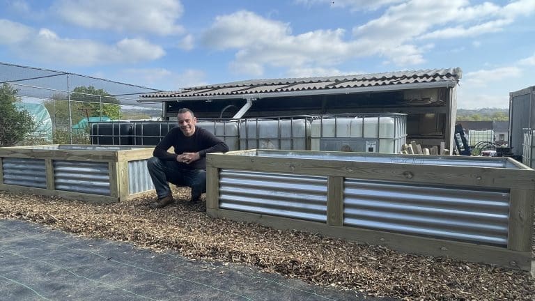 Tony O'Neill crouching beside two large DIY raised beds built from corrugated tin sheets and decking boards in his South Wales garden, with IBC water collection tanks visible in the background