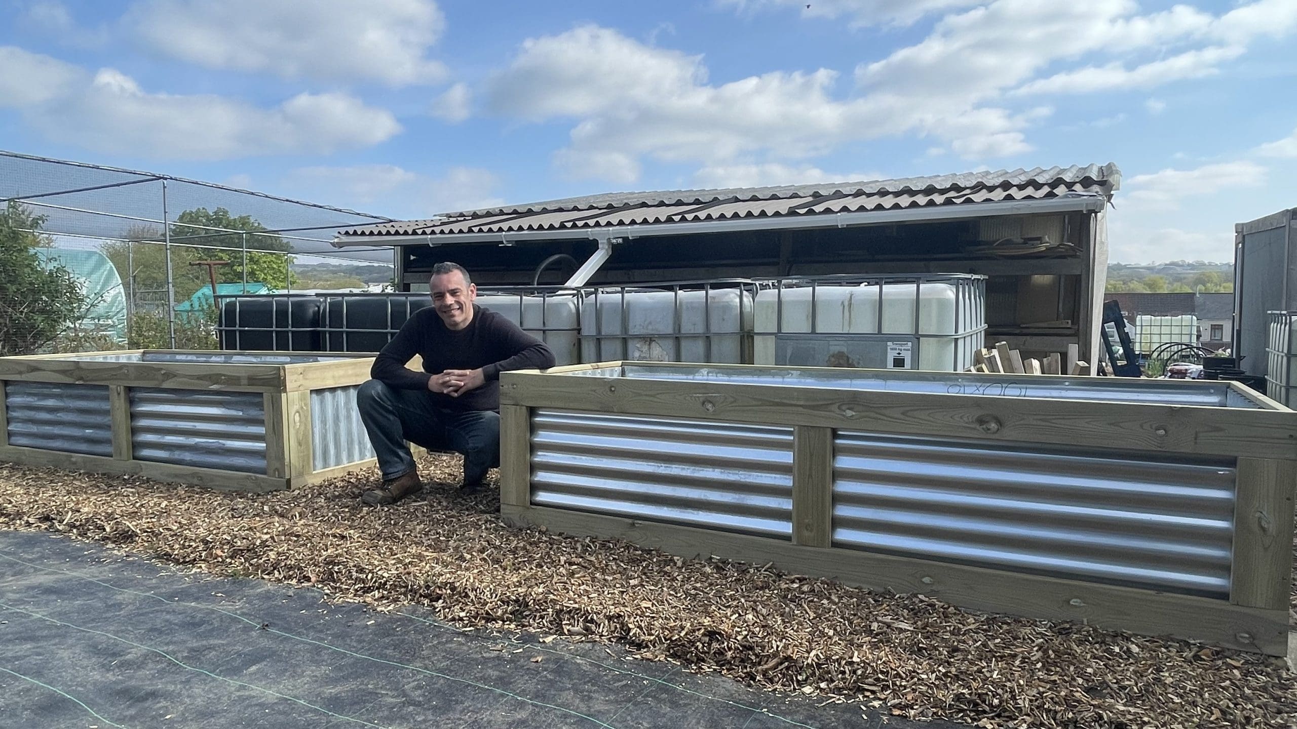 Tony O'Neill crouching beside two large DIY raised beds built from corrugated tin sheets and decking boards in his South Wales garden, with IBC water collection tanks visible in the background