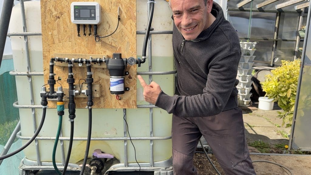 Tony O'Neill pointing to a newly installed Harvst Mate Pro automated irrigation system mounted on an IBC water collection tank in his South Wales garden greenhouse
