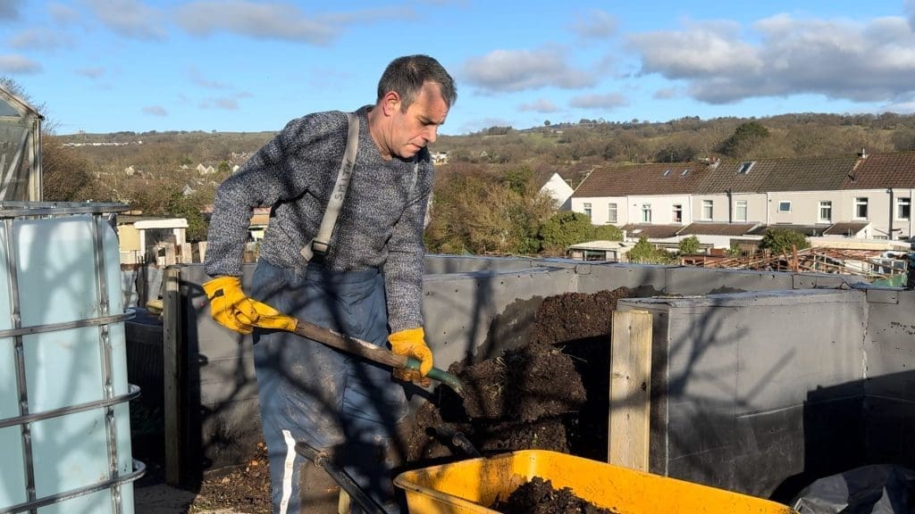 Tony O'Neill wearing yellow gloves and dungarees shovelling dark finished compost from a large bay into a yellow wheelbarrow at his South Wales allotment, with an IBC water tank and South Wales valley visible in the background