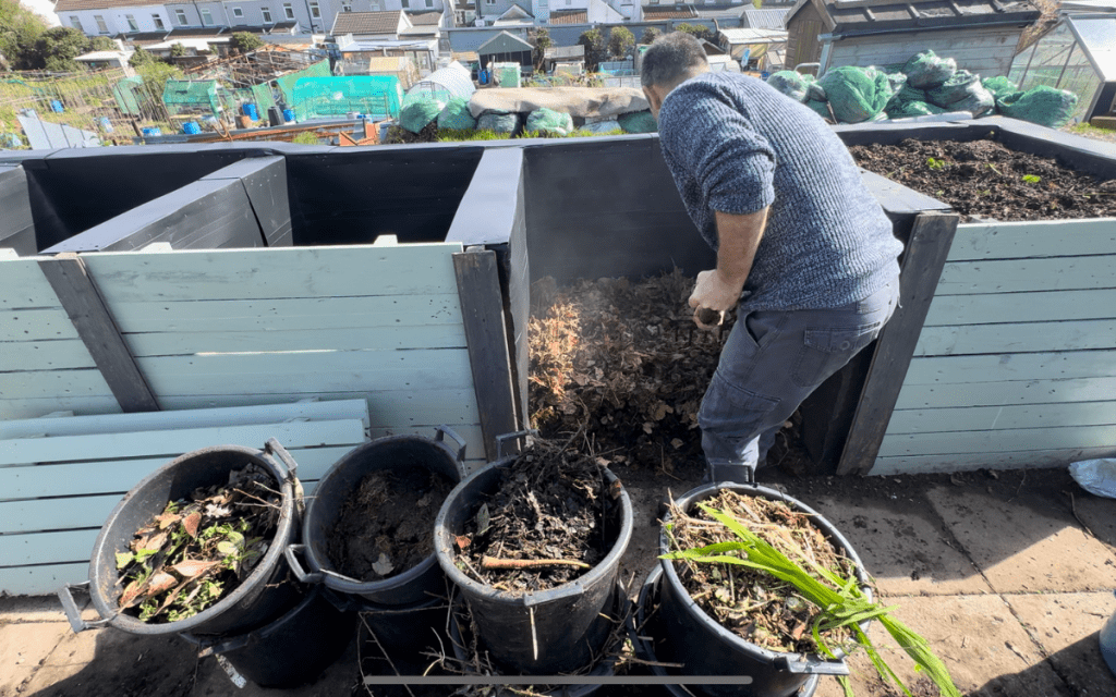 Tony O'Neill turning an active compost bay at his South Wales allotment, with three buckets of fresh green and brown compost material in the foreground and steam rising from the hot compost pile, allotment plots visible in the background